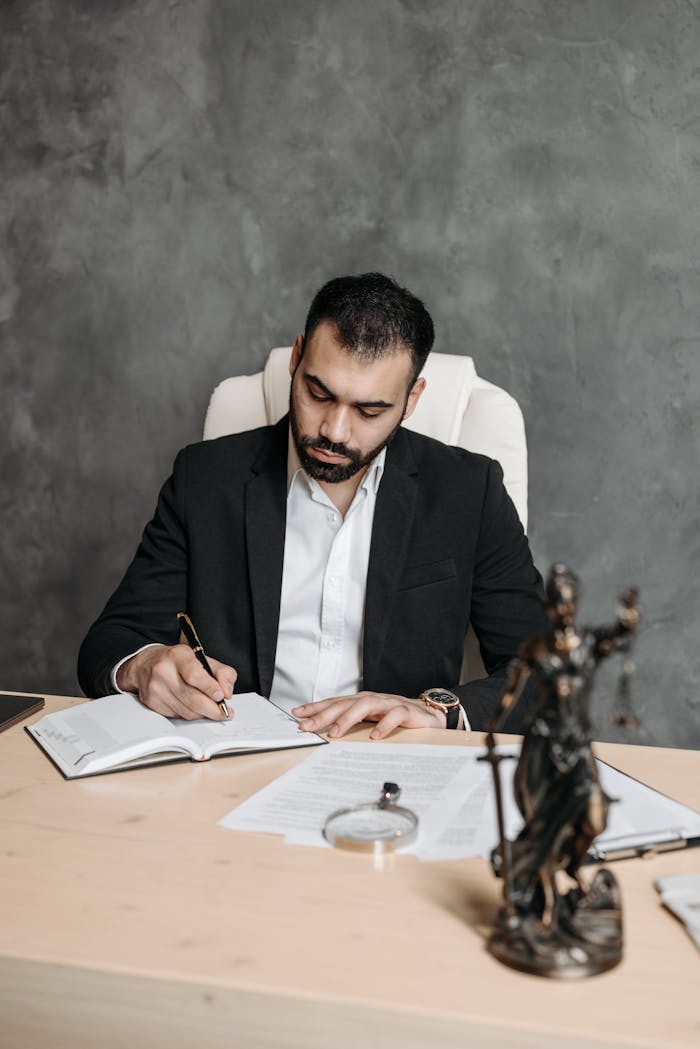 Professional lawyer in a suit taking notes at a desk with legal documents and statue.